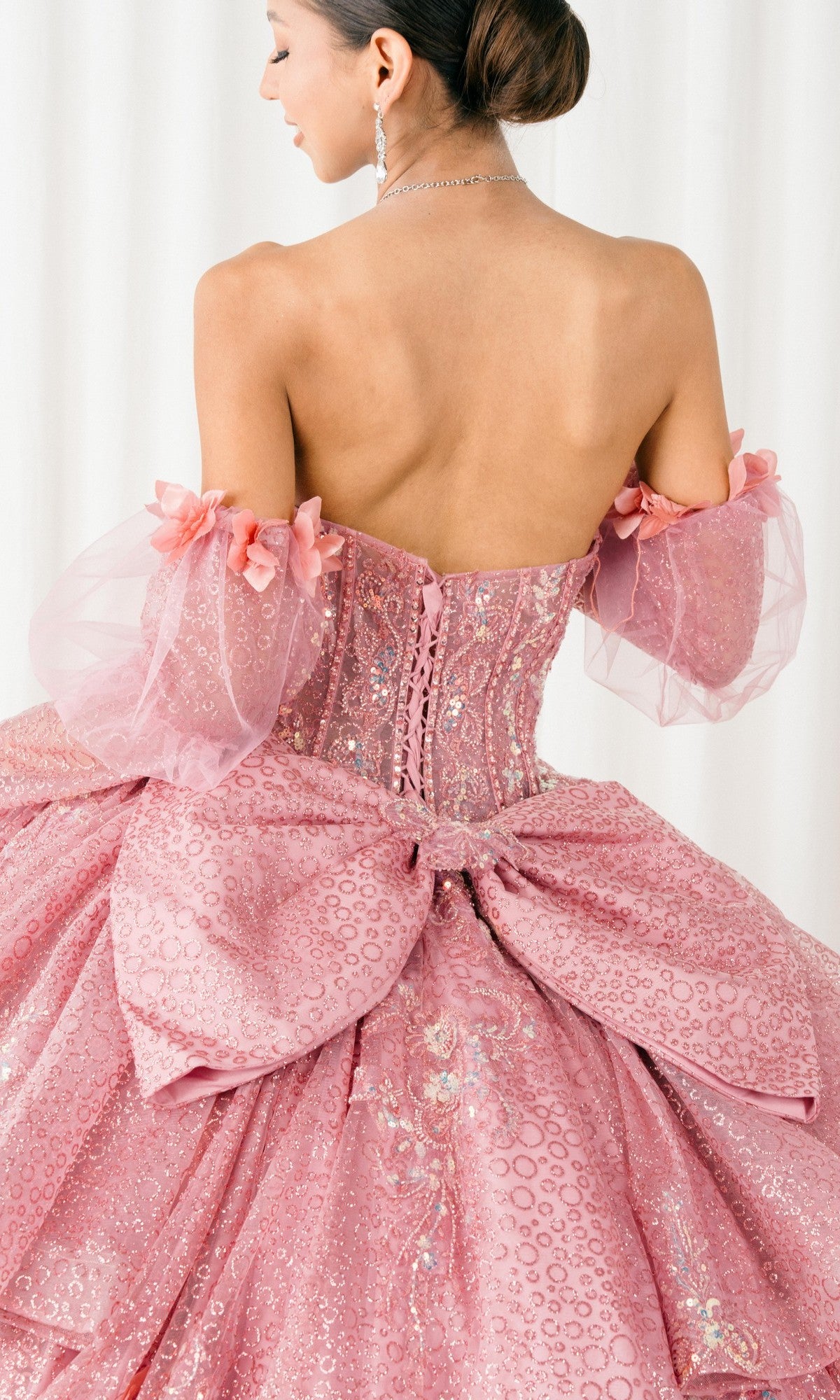 A woman wearing the Quinceanera Dress 8385J By Cinderella Couture, featuring a pink corset bodice, puff sleeves, sequin details, and a large bow, stands against a white background facing away.